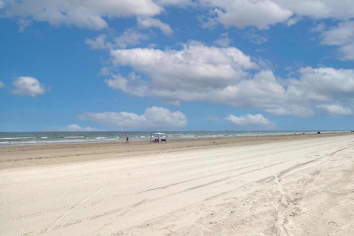 Wide sandy beach stretches along the coast with gentle waves and scattered beachgoers under a partly cloudy sky.