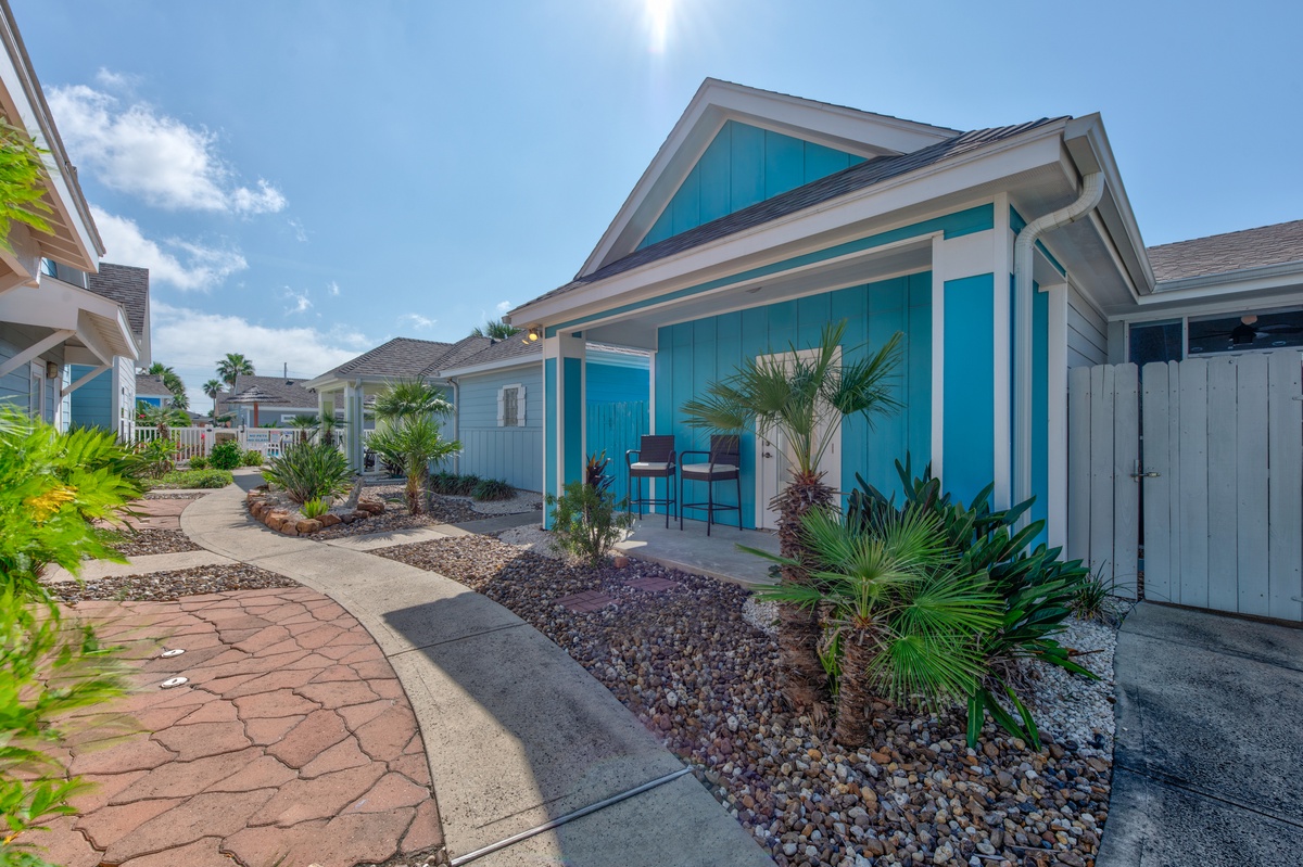 Bright turquoise cottage with tropical landscaping and covered patio seating in a resort-style neighborhood setting.