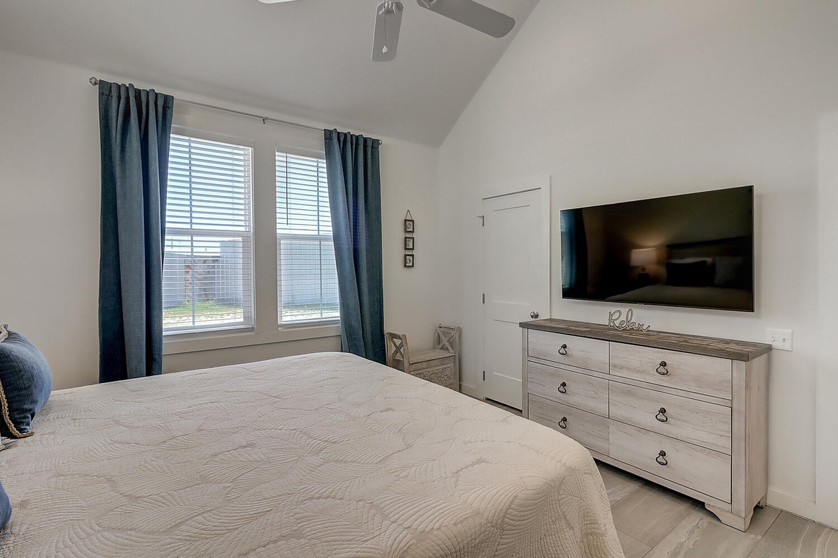 Relax in your serene bedroom with plush bedding, modern TV, and abundant natural light streaming through elegant blue curtains.