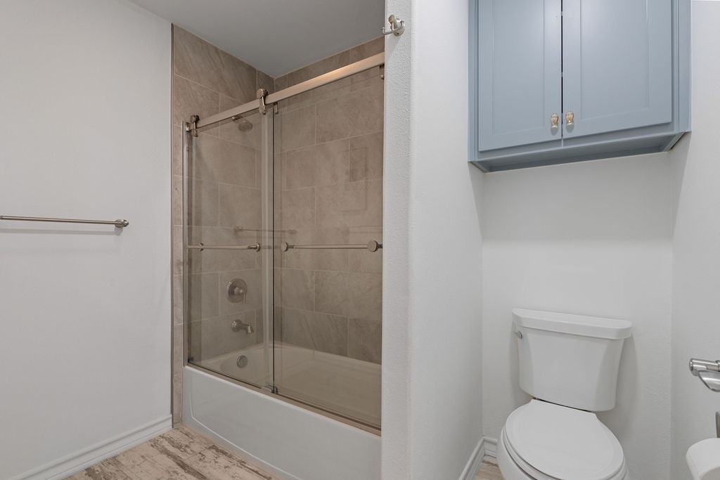 Refresh in your modern bathroom featuring a sleek tub-shower combo with glass doors and thoughtful storage above the toilet for all your essentials.