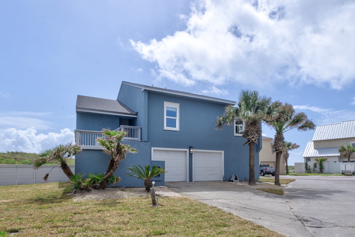 Modern blue beach house with double garage and tropical palm landscaping in coastal neighborhood setting.