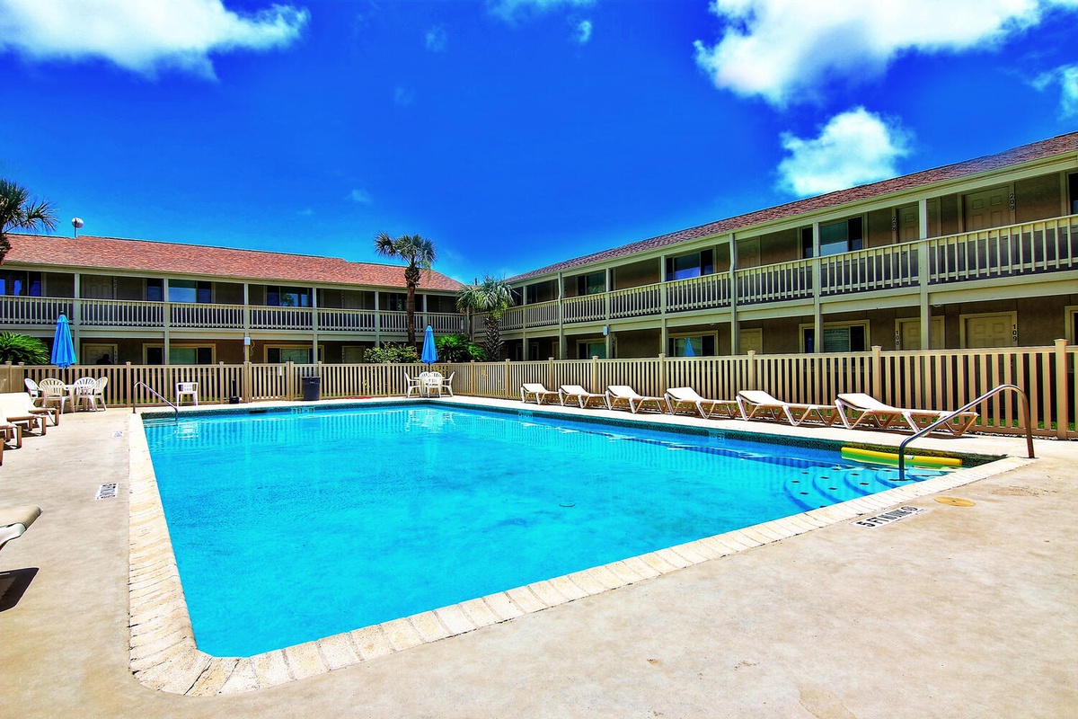 Two-story property buildings surround a sparkling blue swimming pool with lounge chairs and tropical palm trees under bright sunny skies.