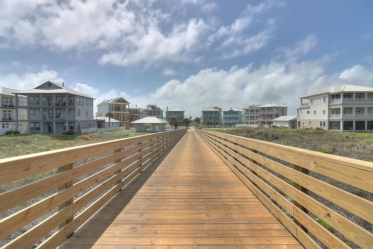 A wooden boardwalk leads through a coastal neighborhood of beach houses under dramatic cloudy skies.