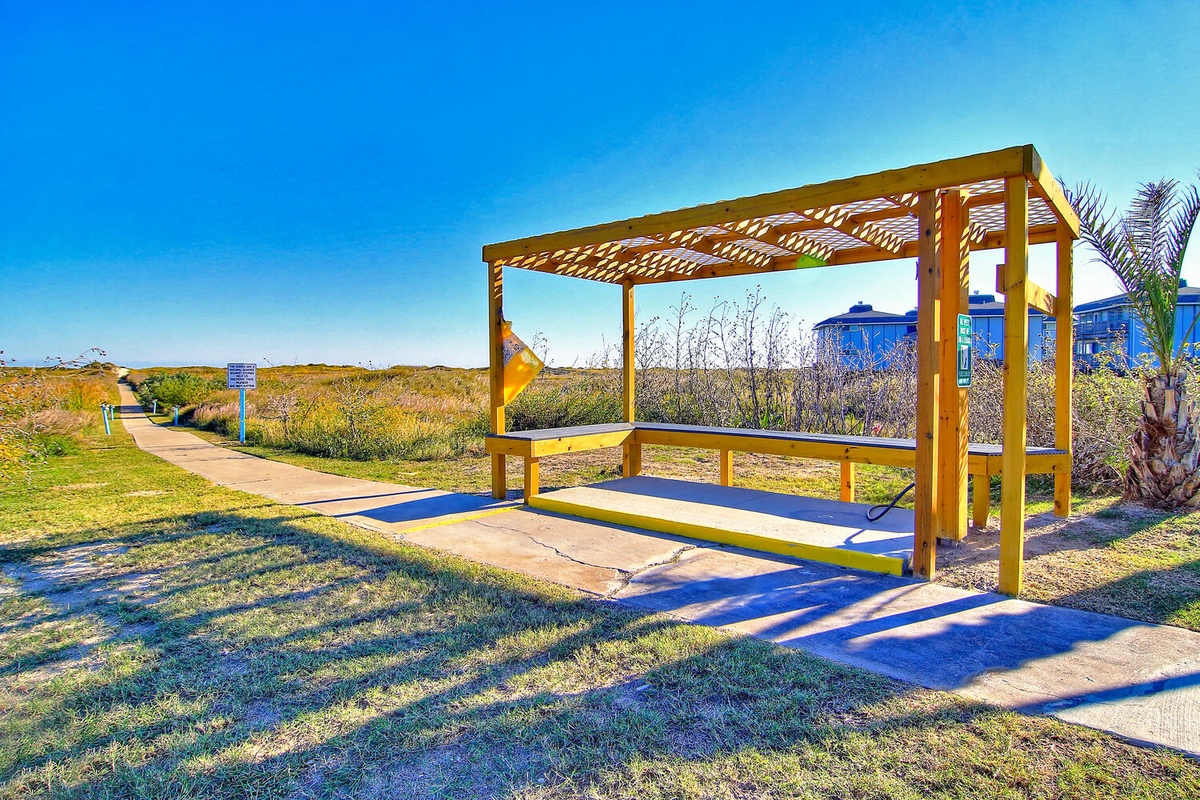 A modern outdoor exercise station sits along a scenic walking trail, surrounded by coastal vegetation and blue skies.