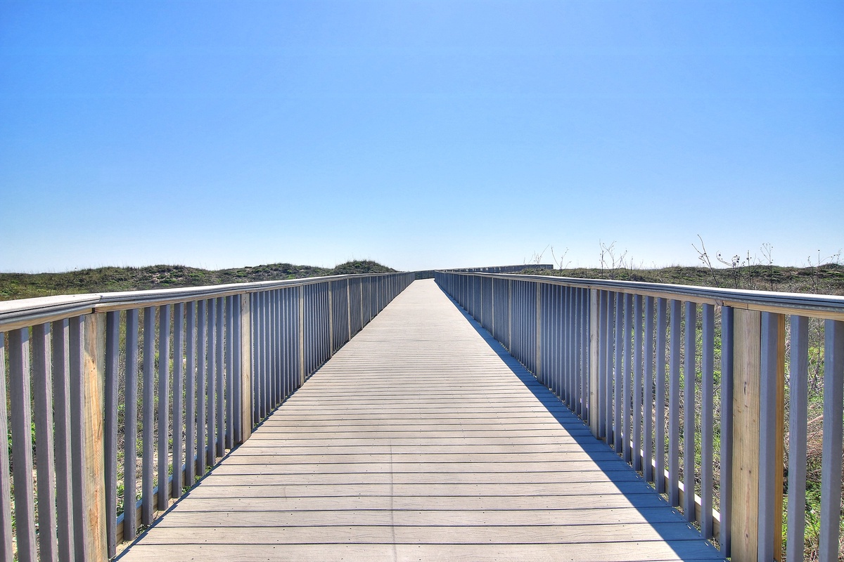 Wooden boardwalk extends through coastal dunes under clear blue skies, providing scenic access to nearby natural areas.