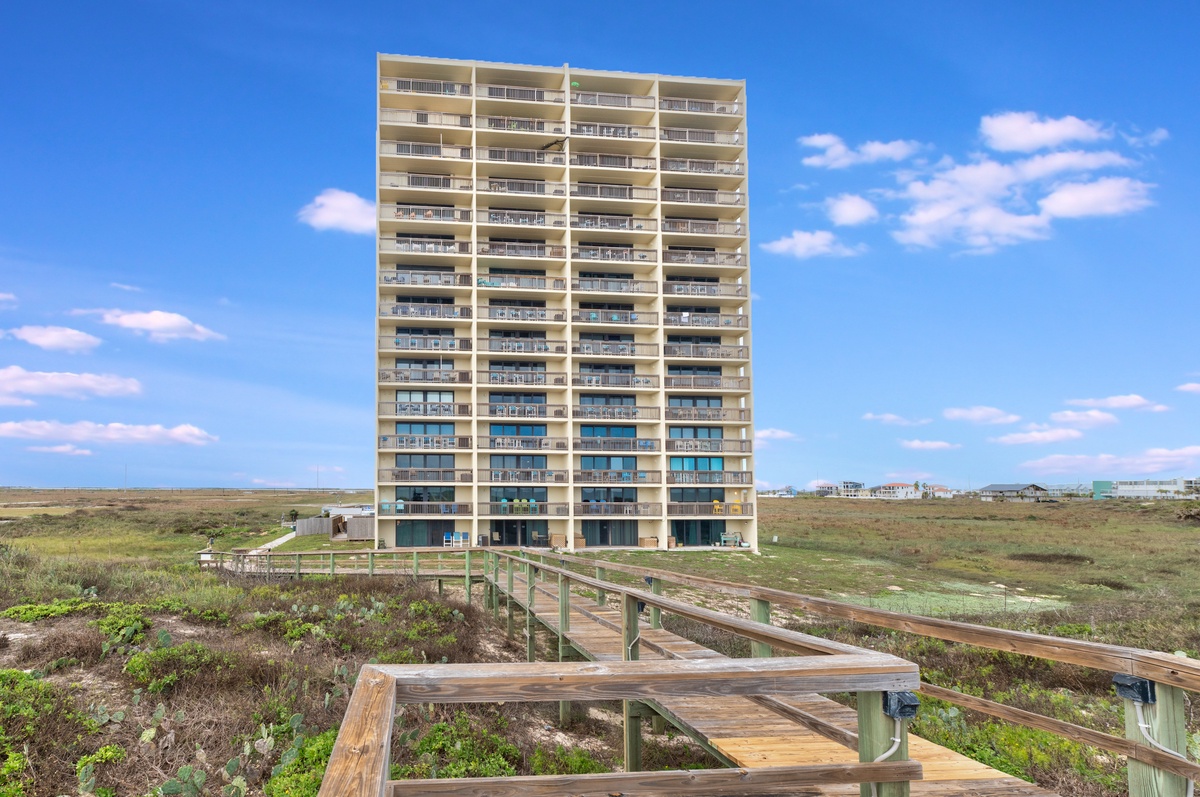 Beachfront high-rise building positioned among coastal dunes and natural vegetation, with elevated walkways providing access through the surrounding landscape.