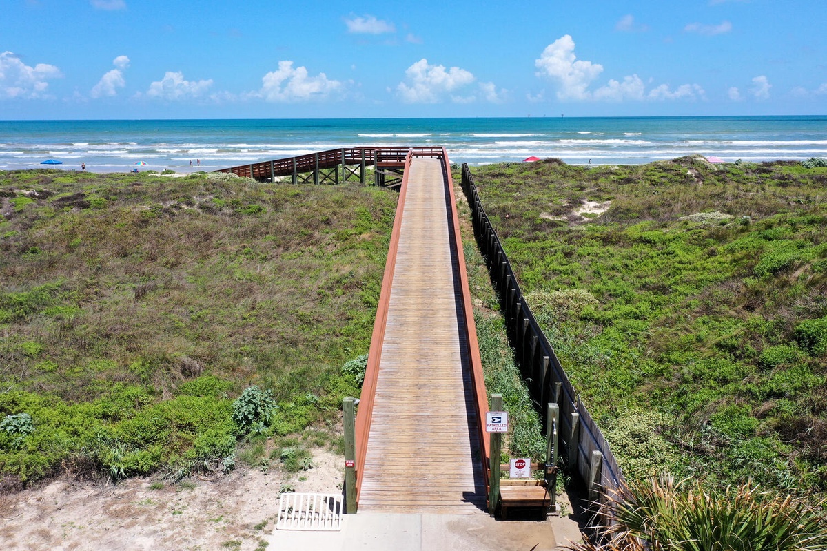 Elevated boardwalk stretches across coastal dunes to pristine beach with rolling waves under brilliant blue skies.