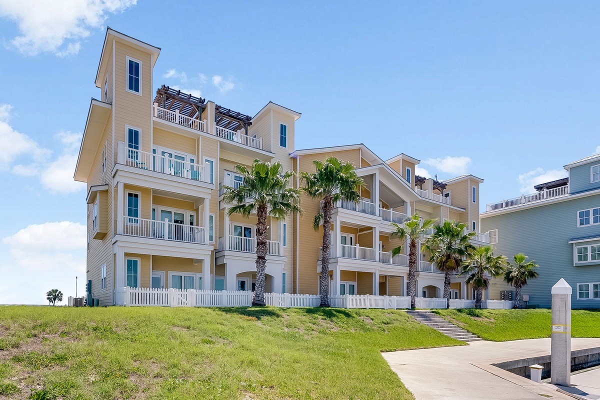 Beachfront resort building features multiple floors with private balconies and tropical palm landscaping under clear blue skies.