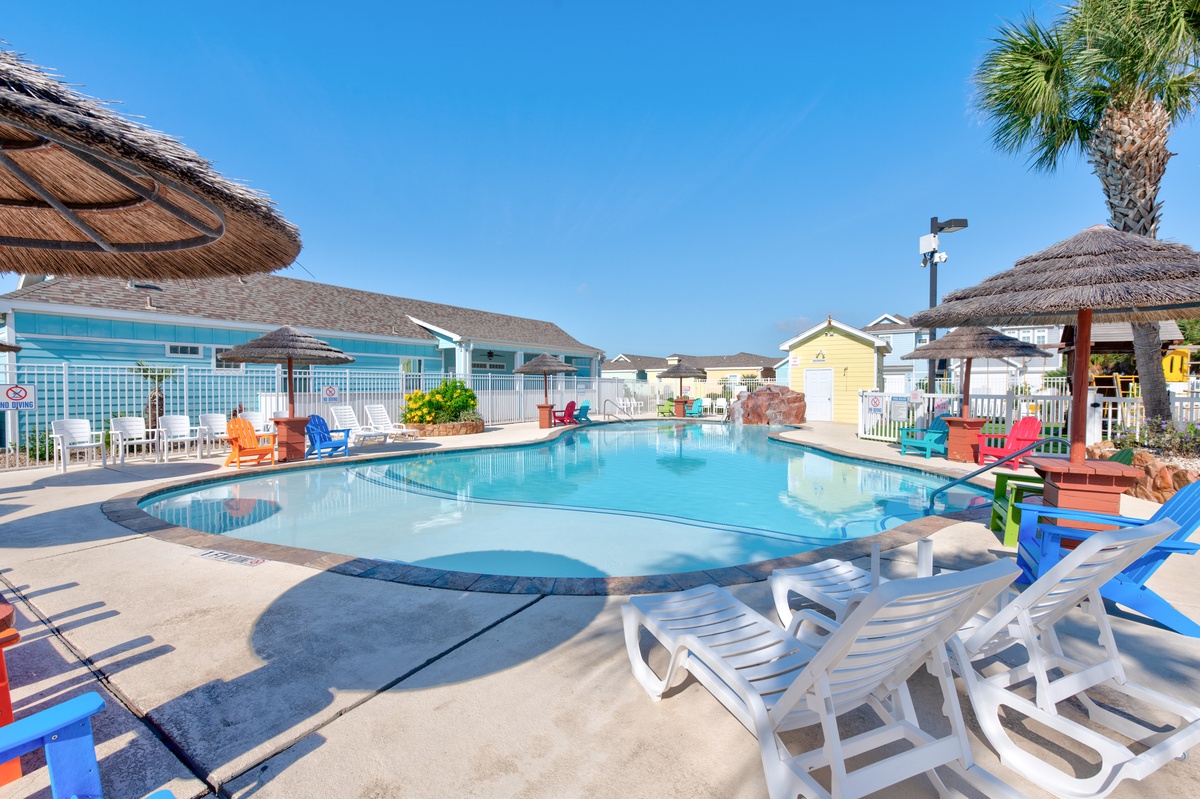 A sparkling resort-style pool area surrounded by colorful beach chairs and tropical thatched umbrellas under clear blue skies.