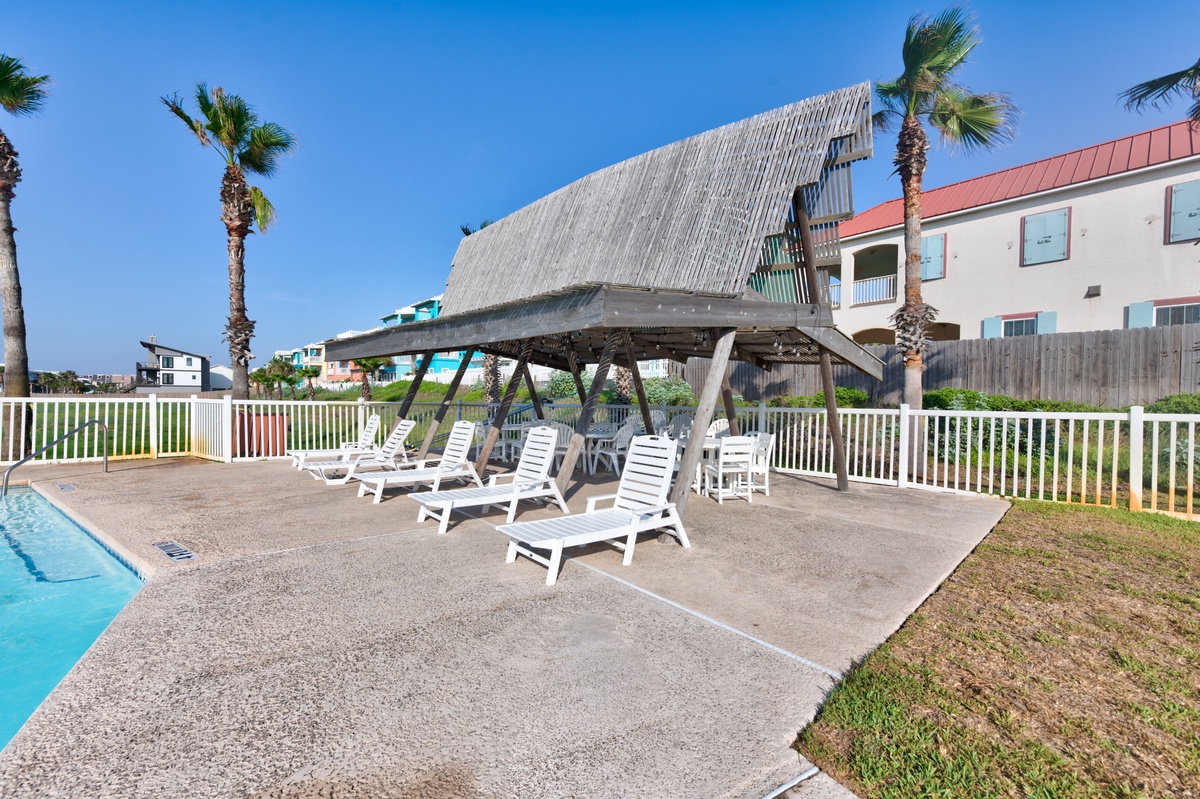 Coastal property features poolside loungers under thatched shade pavilion with palm trees and red-roofed buildings nearby.
