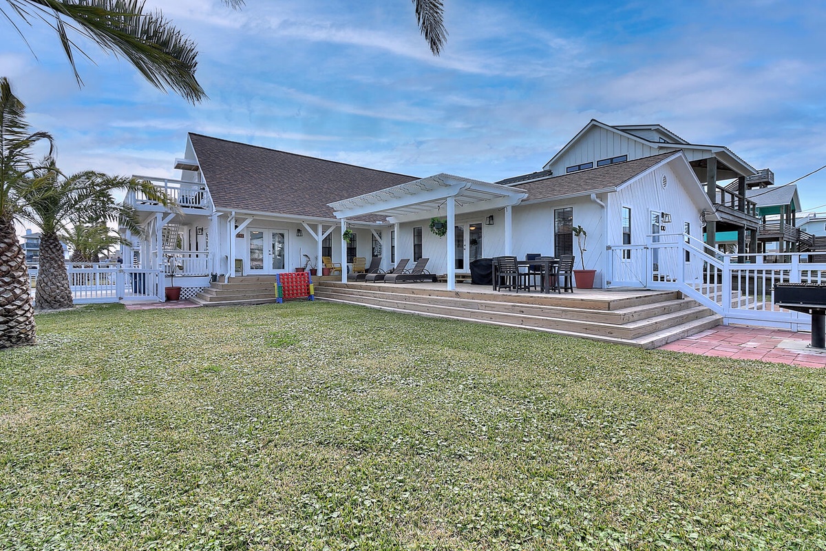 A coastal beach house with wraparound porch and spacious lawn sits under swaying palm trees in bright daylight.