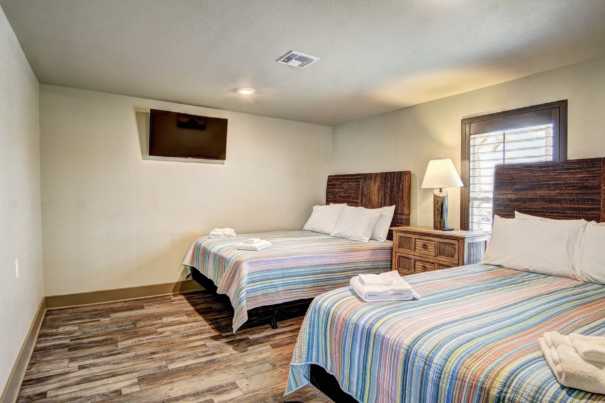 Relax in your comfortable twin bedroom with colorful striped bedding, wicker headboards, and modern amenities including a wall-mounted TV for cozy evenings.