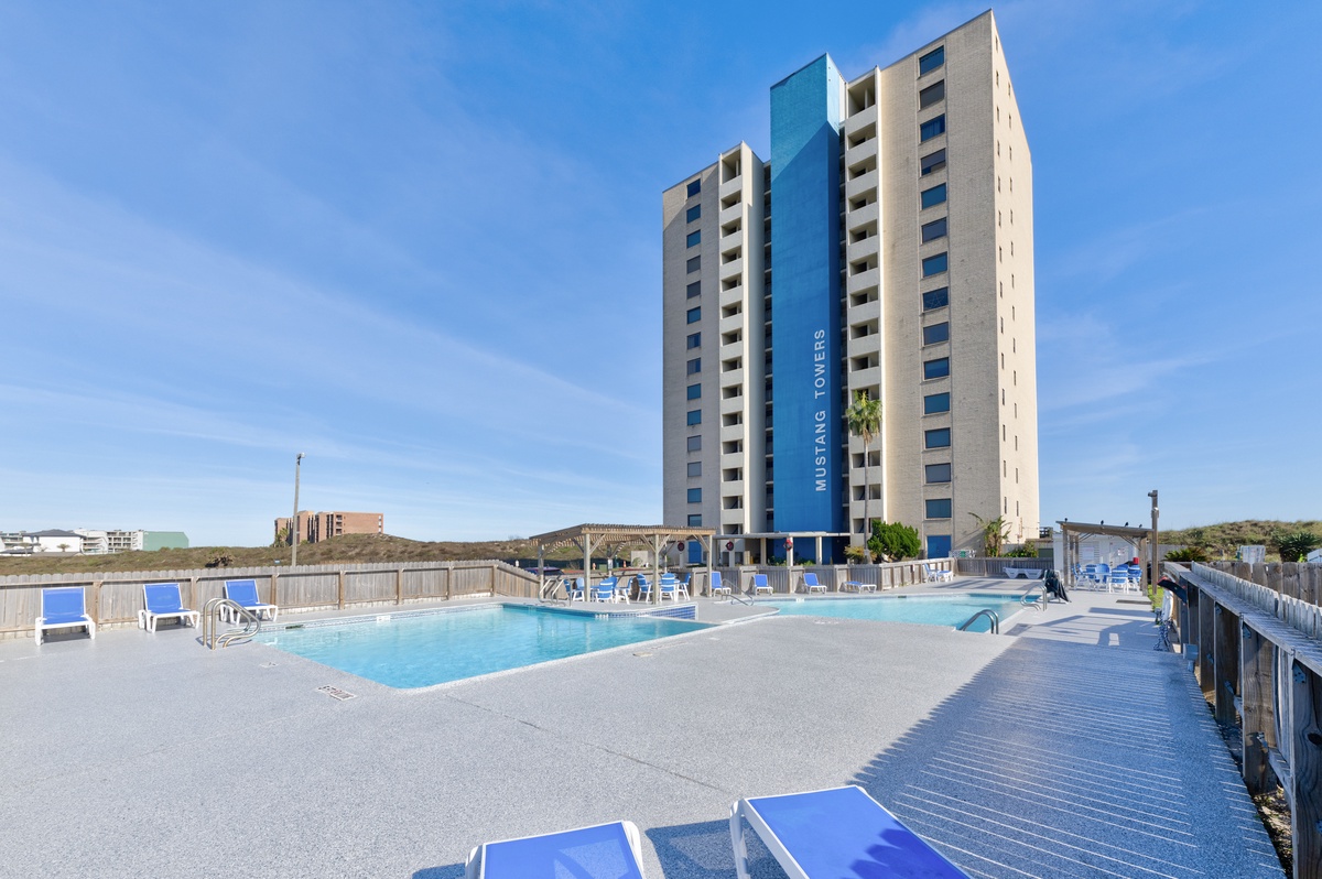 Modern beachfront towers with inviting pool area featuring bright blue loungers under expansive coastal skies.