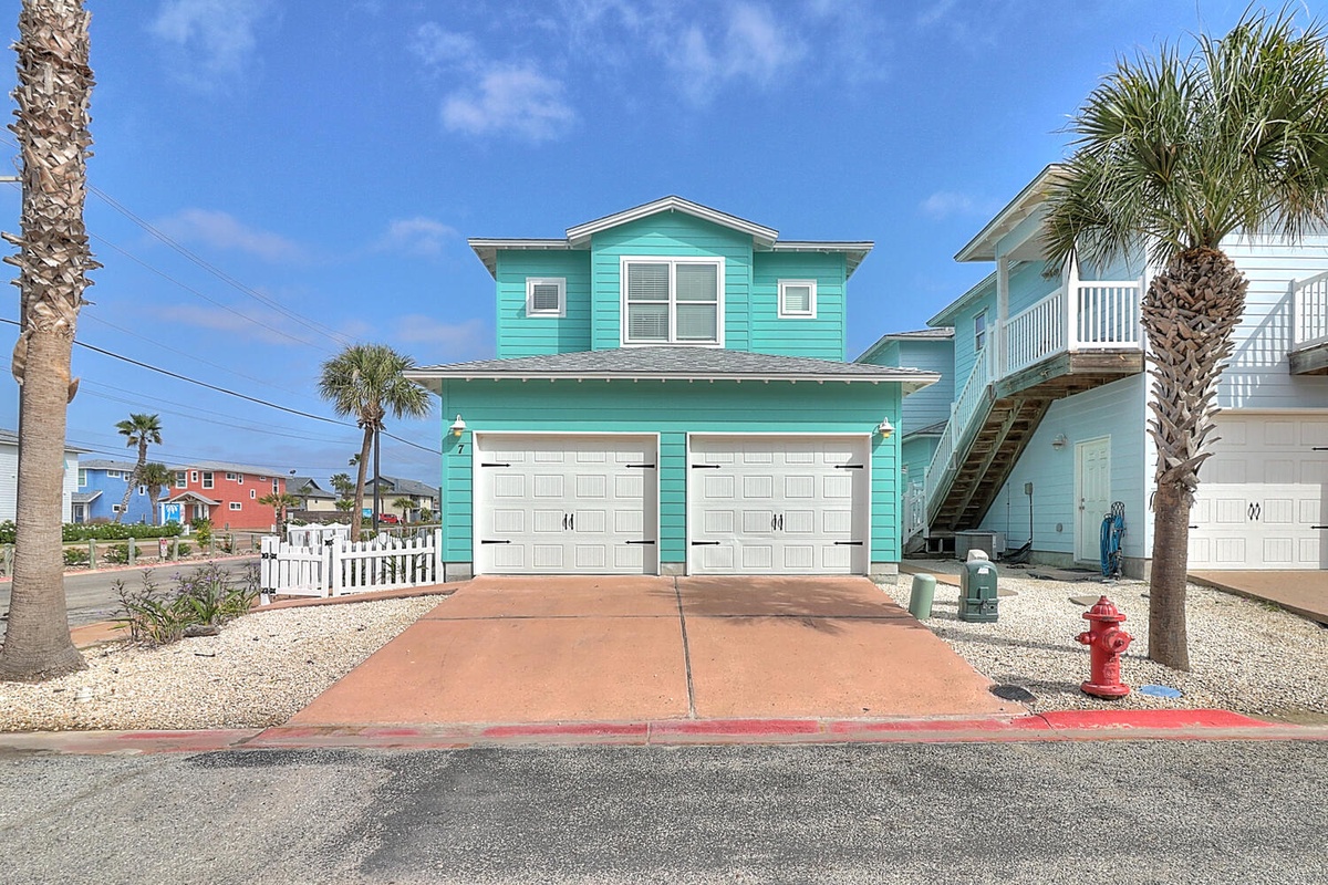 Charming turquoise beach house with double garage and palm trees in a vibrant coastal neighborhood.