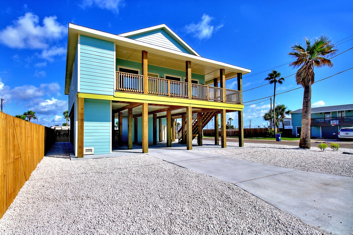 Charming turquoise beach house on stilts with covered parking and tropical palm trees under sunny coastal skies.