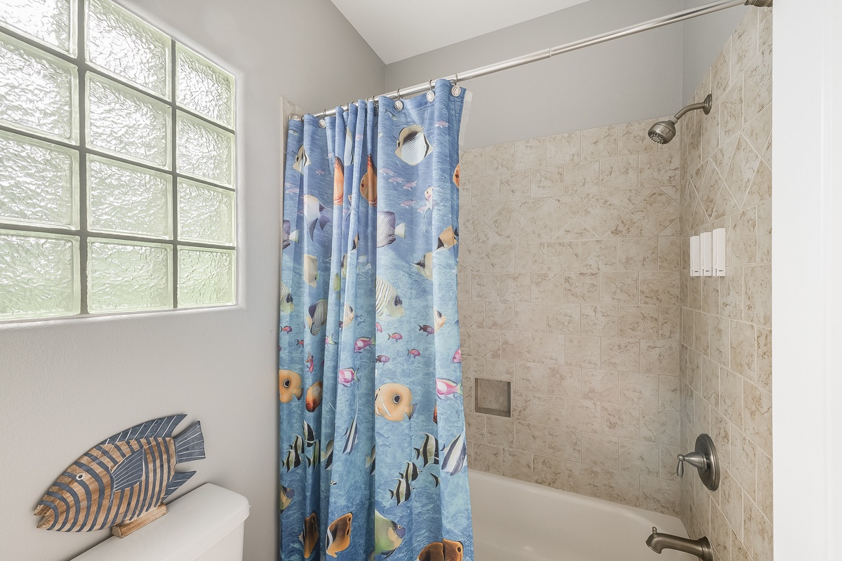Your private bathroom features a soothing tub-shower combo with playful ocean-themed curtain, natural light, and neutral tile design.
