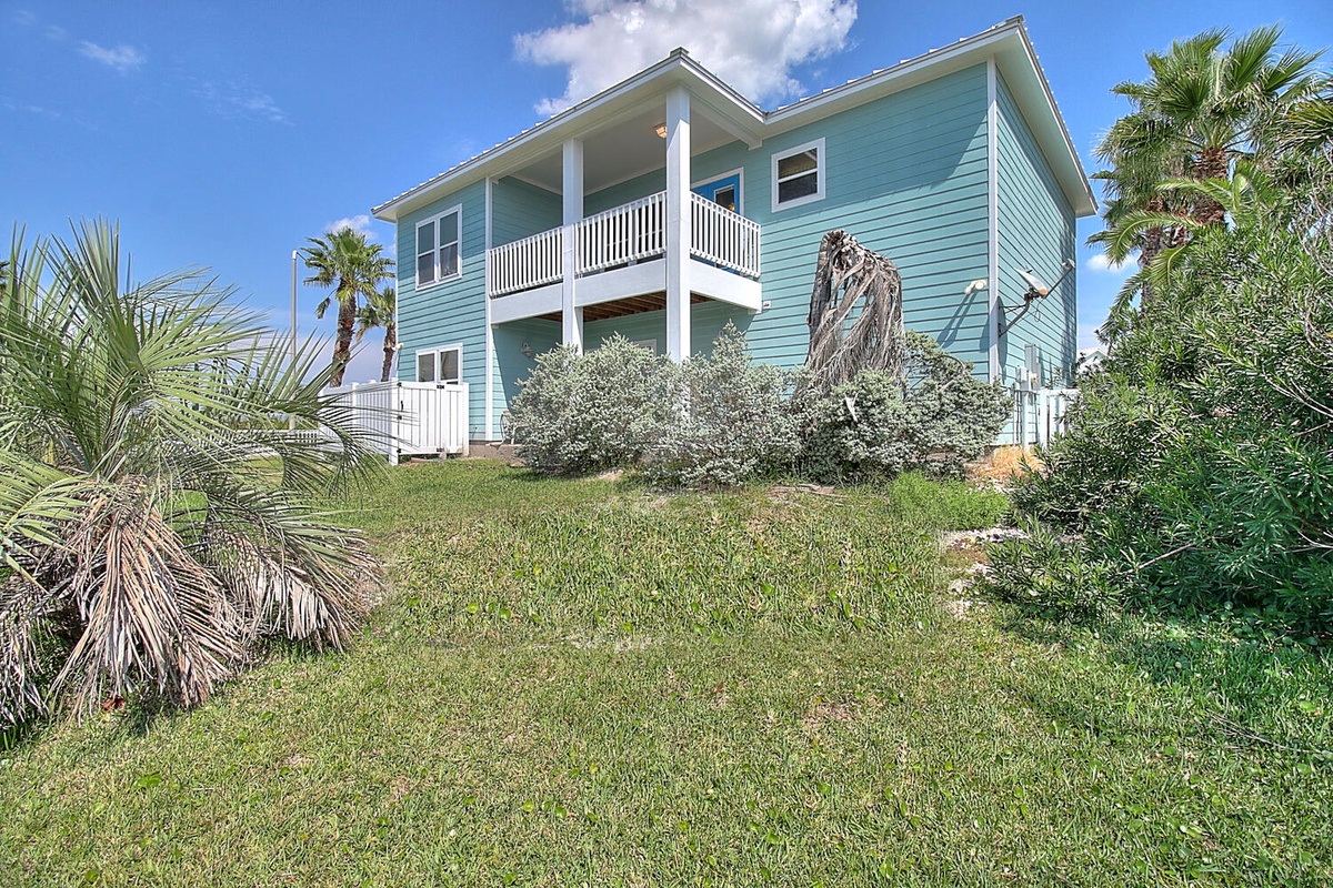 Coastal two-story property featuring turquoise siding, white balcony railings, and lush tropical landscaping with palm trees surrounding the building.