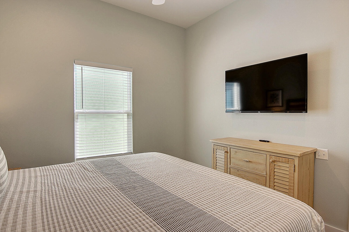 Relax in your peaceful bedroom with cozy linens, wall-mounted TV for entertainment, and natural light streaming through blinds.