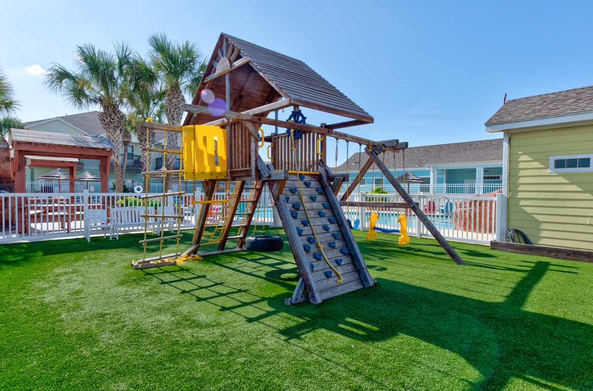 Colorful beachside resort with playground featuring swings, climbing wall, and slide surrounded by palm trees and vacation cottages.