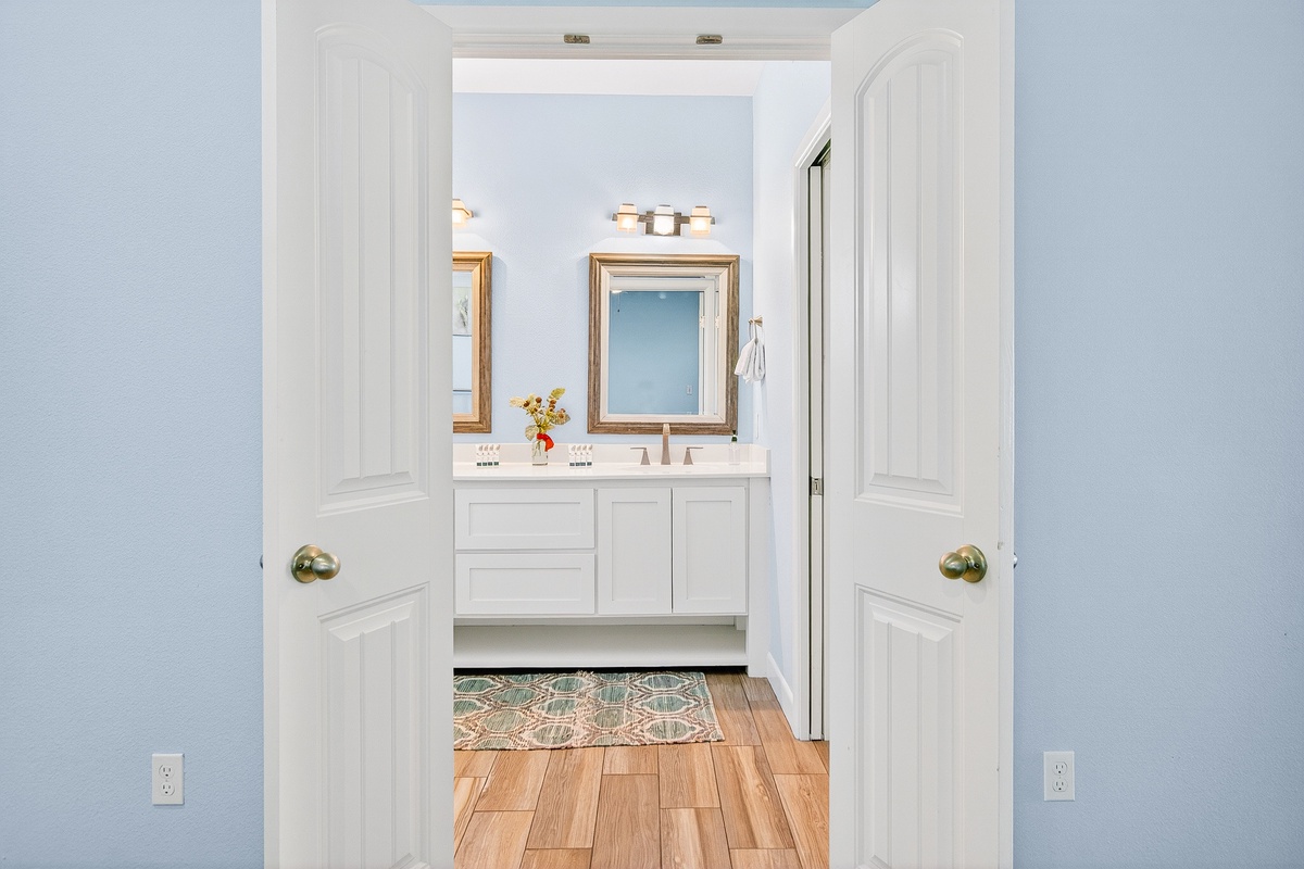 Step into your elegant en-suite bathroom featuring pristine white cabinetry, warm wood floors, and thoughtful lighting that creates a spa-like atmosphere for your morning routine.