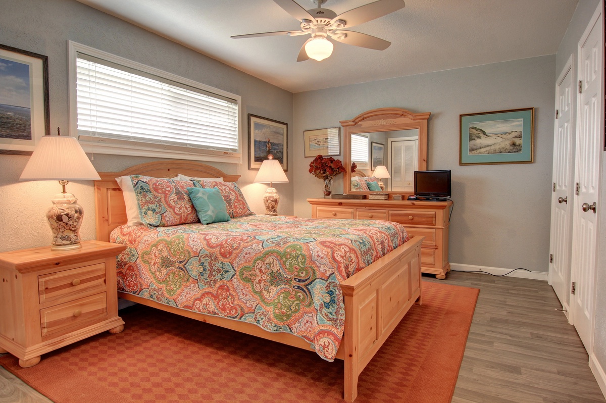 Relax in your beautifully appointed bedroom featuring colorful bedding, warm wood furnishings, and coastal artwork creating a peaceful retreat.