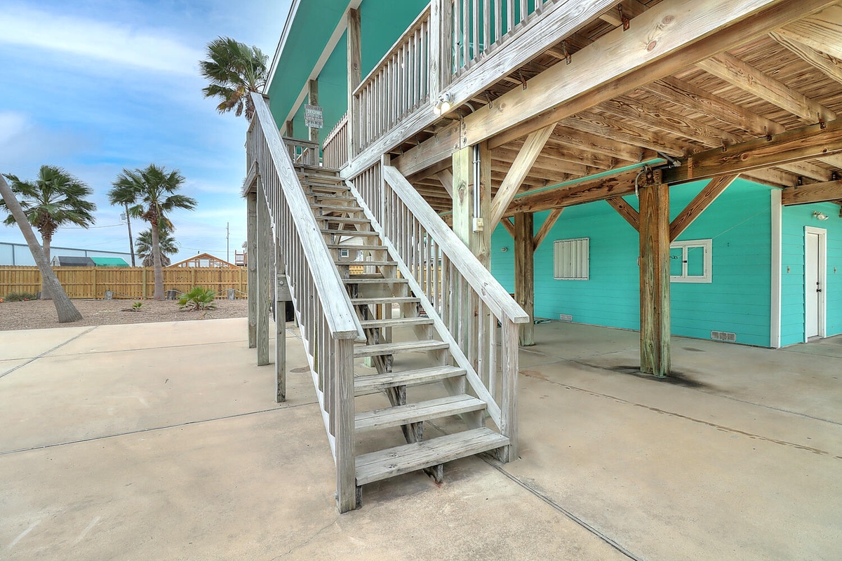Bright turquoise beach house with wooden stairs leading to upper level balcony and palm trees swaying nearby.