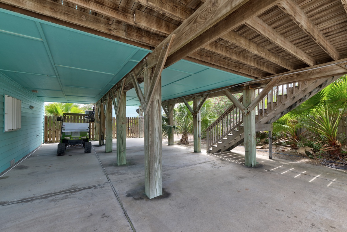 Protected parking beneath the main house, with sturdy timber beams and tropical landscaping framing the covered area. A wooden staircase leads to the elevated living space above.
