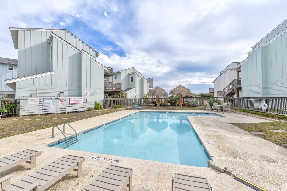 Shared pool area surrounded by coastal vacation homes with tropical tiki huts in the background.