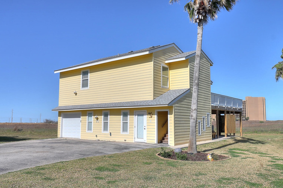 Bright yellow vacation home with white garage and tropical palm tree, situated in peaceful coastal setting.