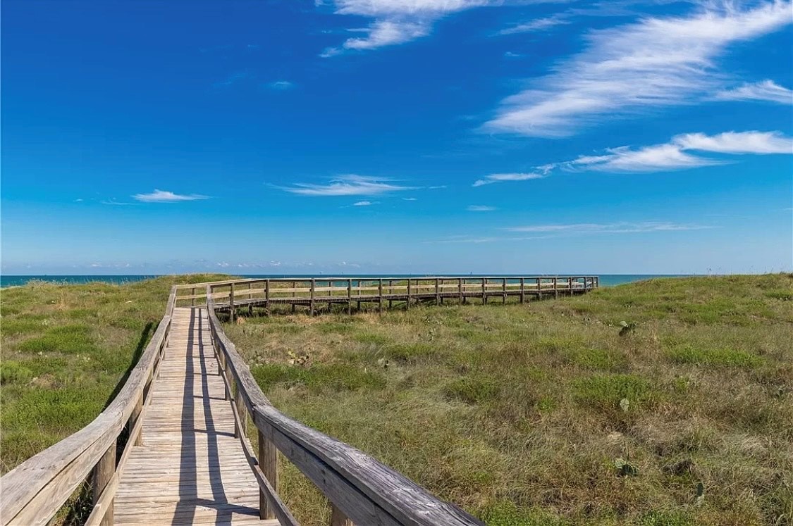 Wooden boardwalk leads through coastal dunes toward the ocean under brilliant blue skies with wispy clouds.