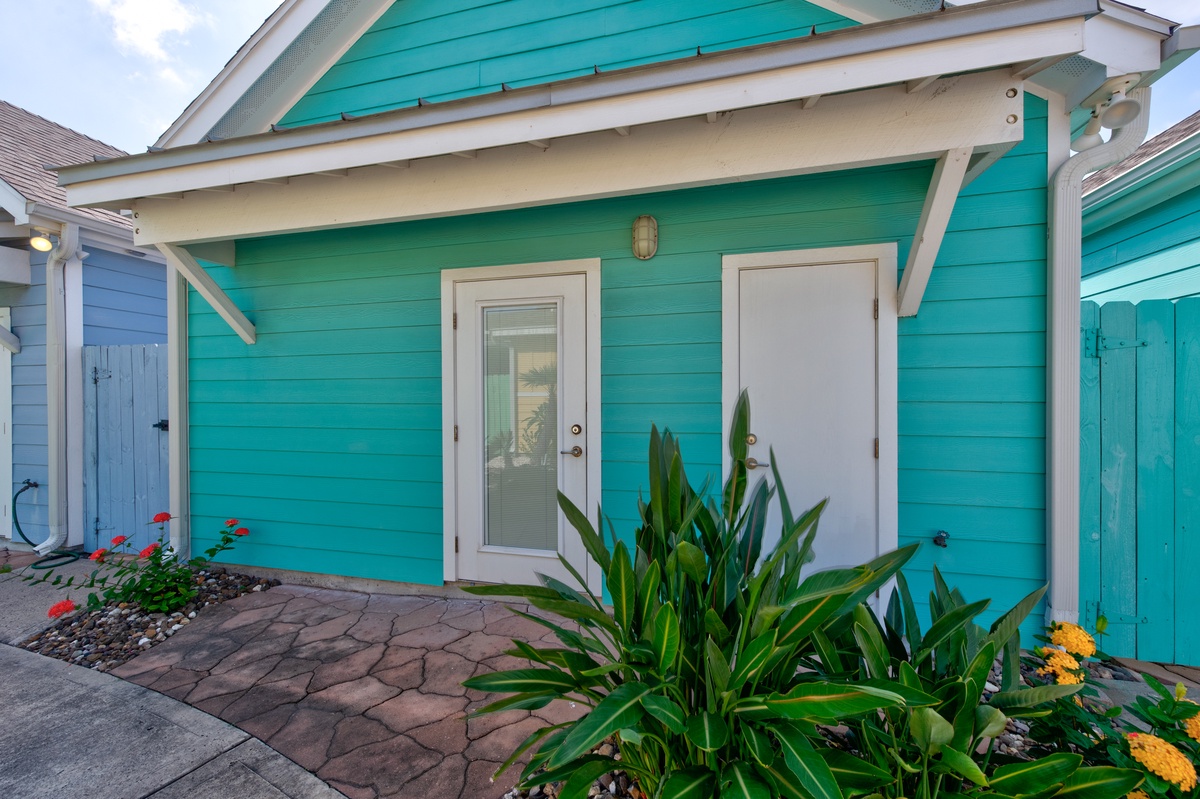 Charming turquoise cottage entrance welcomes guests with vibrant tropical plants and cheerful coastal architecture.