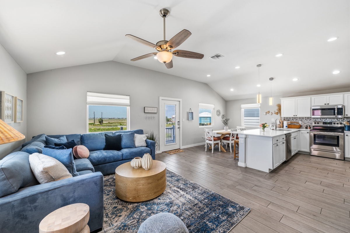 Unwind in your spacious open-plan living area with vaulted ceilings, cozy blue sectional, and gleaming kitchen island—all bathed in natural light from windows overlooking serene coastal marshlands.