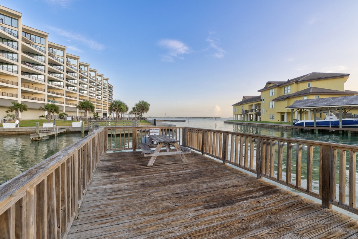 Waterfront pier with picnic table overlooking calm bay, flanked by multi-story condominiums and palm trees—perfect for peaceful mornings or sunset watching in a scenic coastal setting.