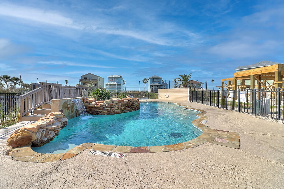 Resort-style pool with waterfall feature and coastal property views under sunny blue skies.