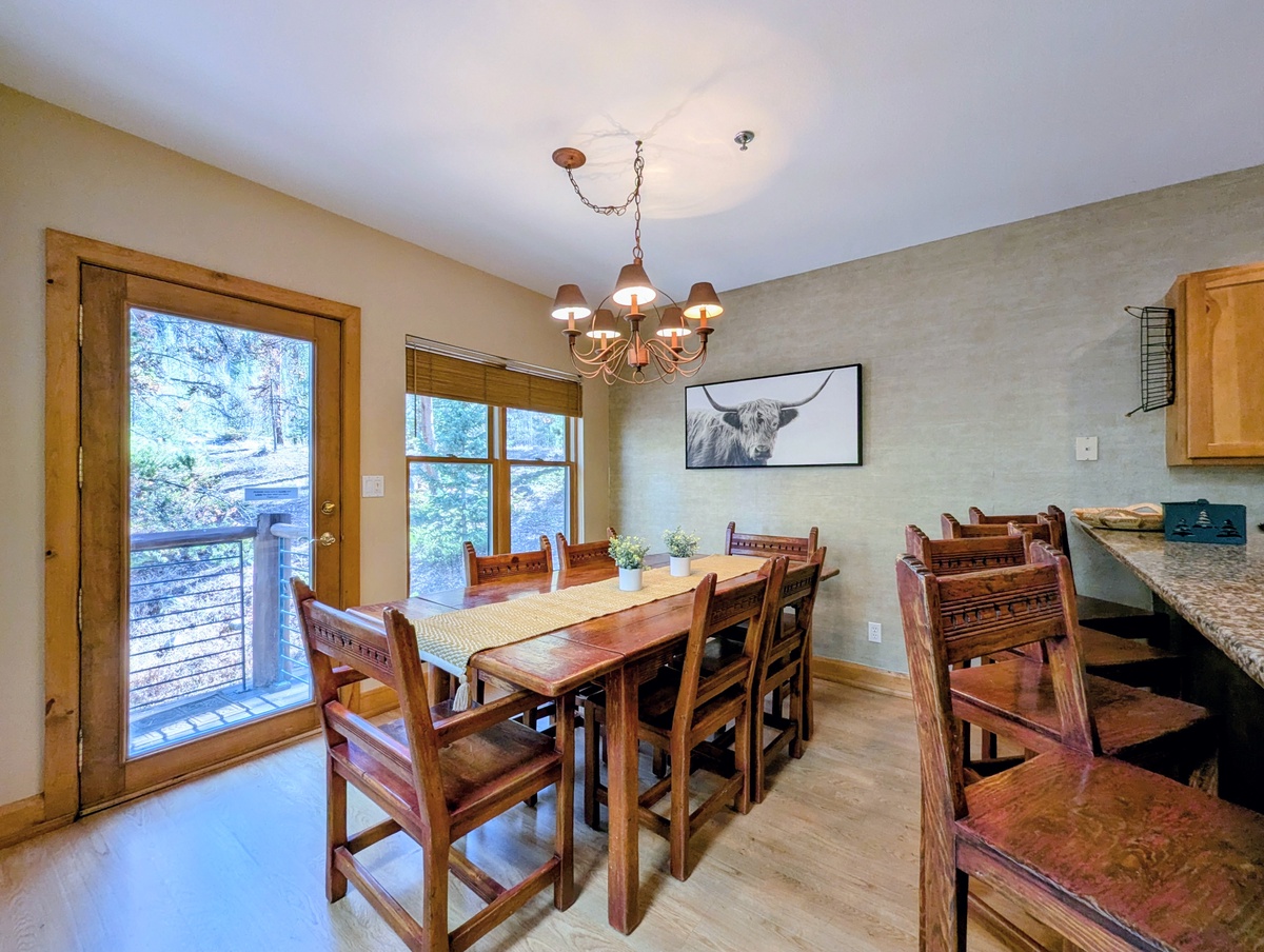 Gather around your rustic dining table where warm wood tones and charming Highland cattle artwork create an inviting atmosphere for shared meals. Natural light pours through the deck door, connecting you to the peaceful forest views beyond.