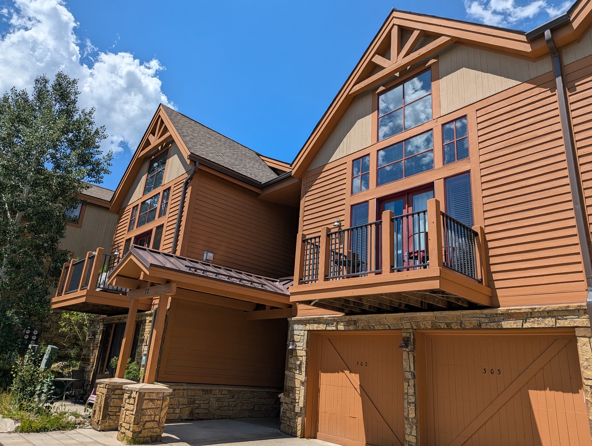 A striking mountain lodge-style residence with warm wood siding, expansive windows, and scenic balconies set against a bright blue sky.