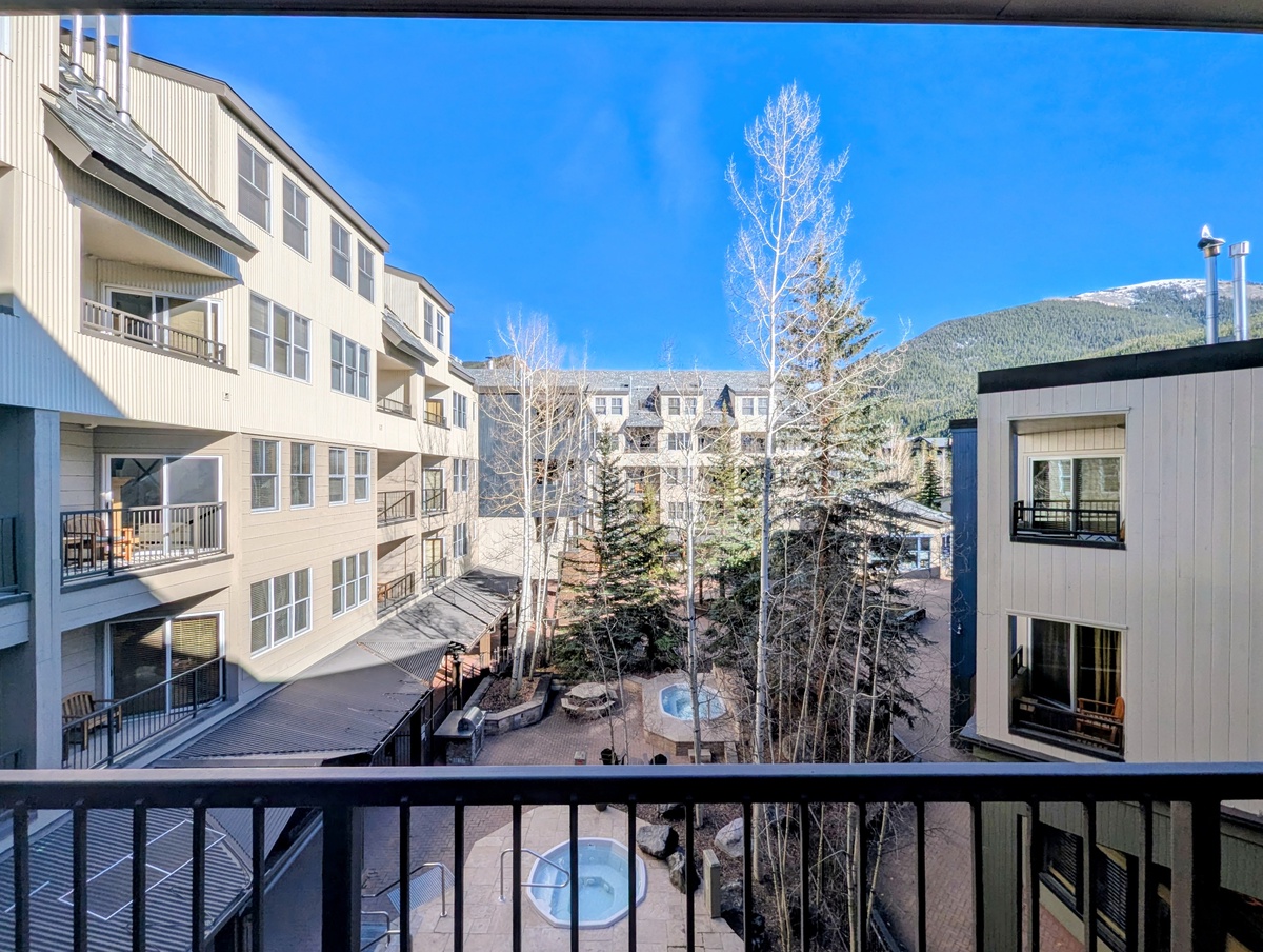 A mountain community unfolds beneath clear blue skies, with forested peaks rising beyond residential buildings and courtyard spaces dusted with early winter snow.