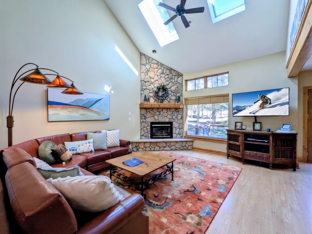 Gather in this sunlit living room with soaring vaulted ceilings, a striking stone fireplace, and plush leather seating. Natural light pours in from skylights and windows, inviting you to unwind after your mountain adventures.