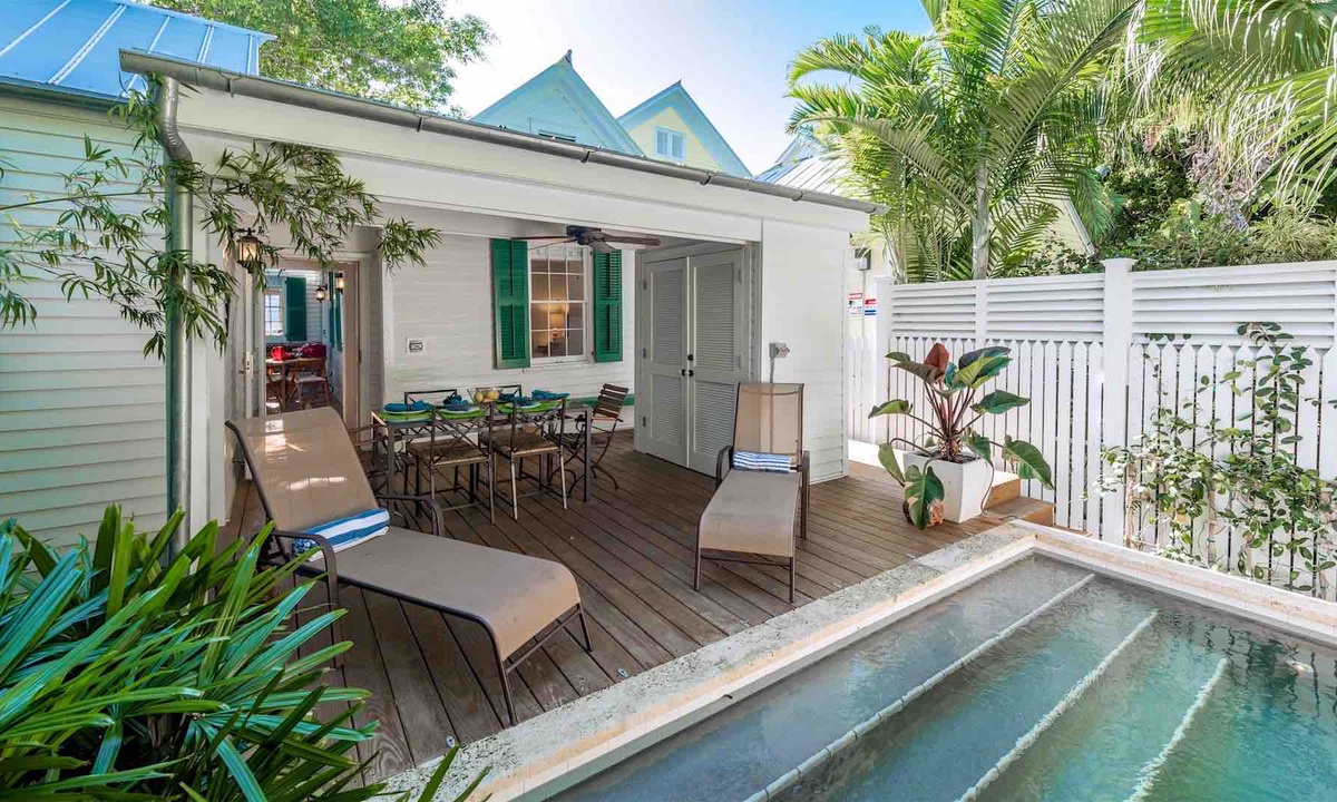 The back porch of a Key West vacation rental. An outdoor table with four chairs and two loungers leads up to the steps of an outdoor pool.