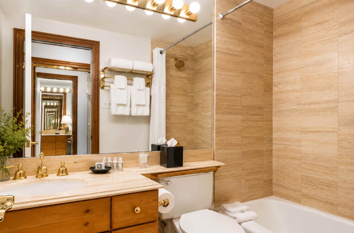 Sink into your private bathroom's warm elegance, where golden fixtures and soft lighting create a spa-like retreat. Fresh towels await, while wood accents add natural charm to your daily routine.