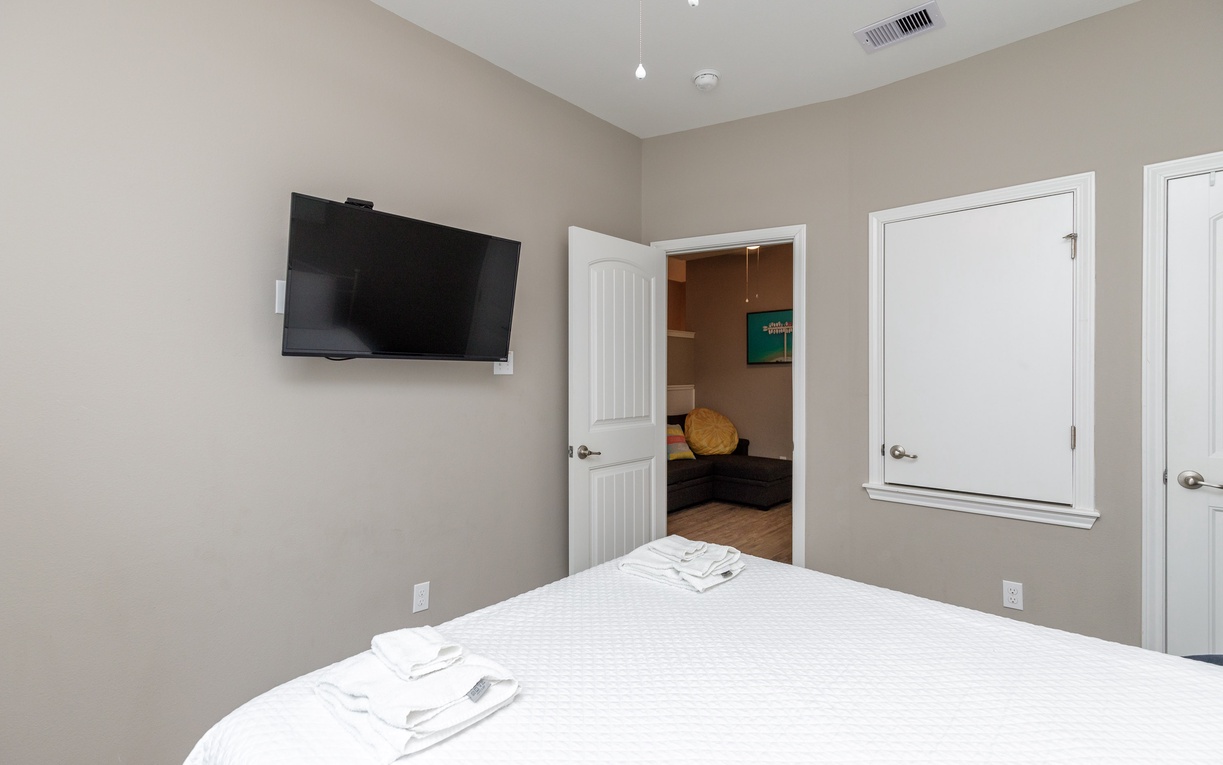 Your peaceful bedroom features a wall-mounted TV and fresh towels, with an inviting glimpse into the connected living space.
