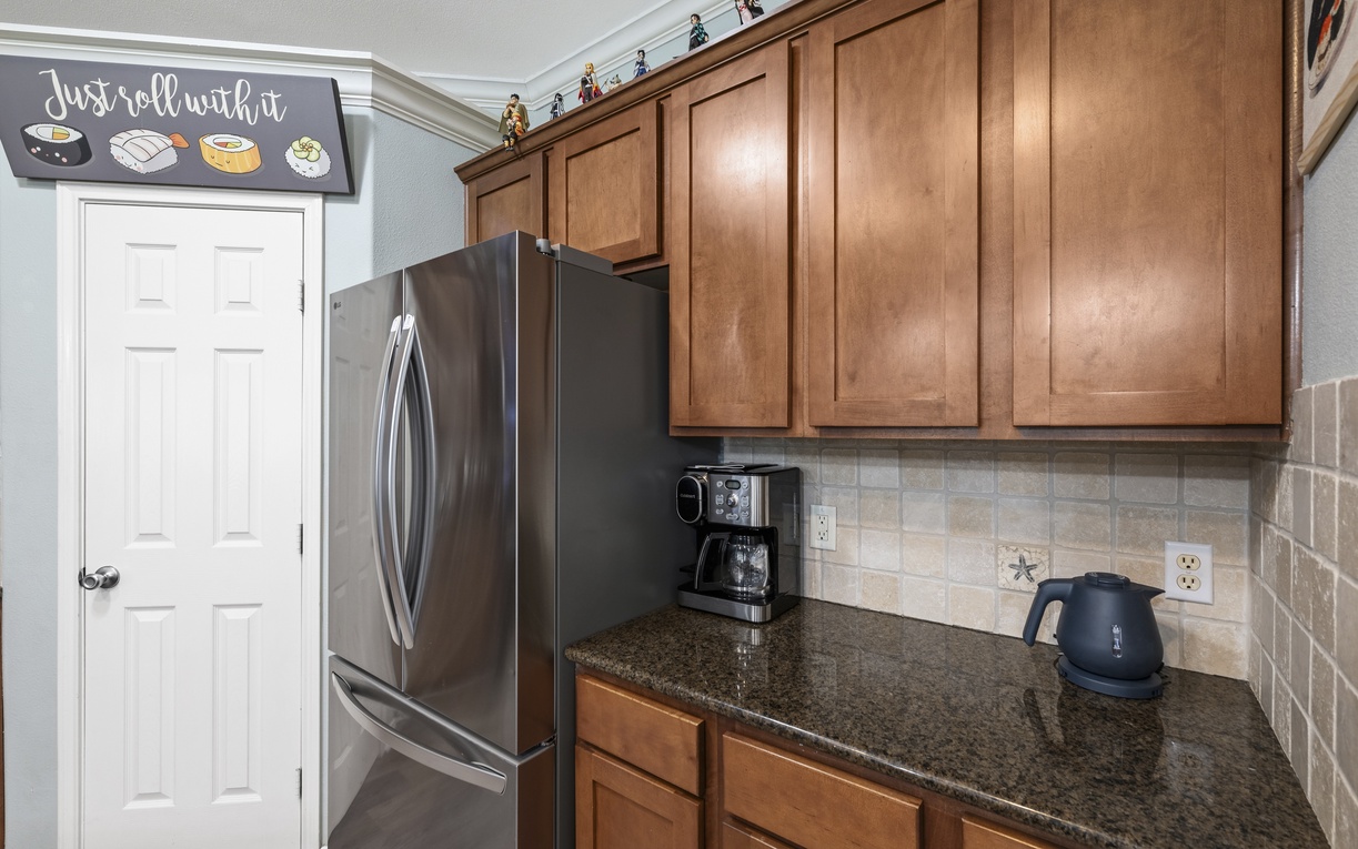 Your morning coffee station awaits with a stainless steel refrigerator, rich wood cabinetry, and modern appliances on sleek granite countertops—everything you need for a cozy start.