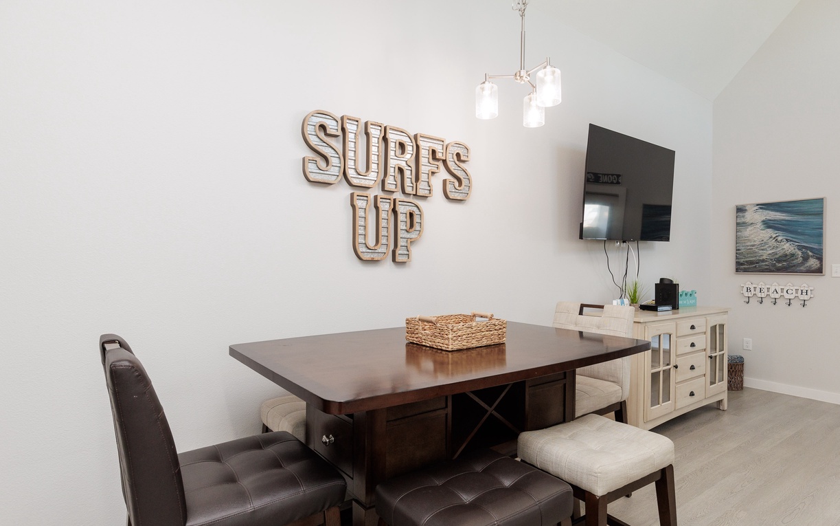 Your stylish dining space features coastal charm with 'SURF'S UP' wall art and a warm wood table perfect for family meals.