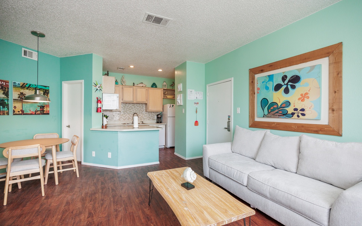 Relax in your bright, coastal-themed living space with cheerful turquoise walls, comfortable seating, and an open kitchen perfect for preparing meals together.
