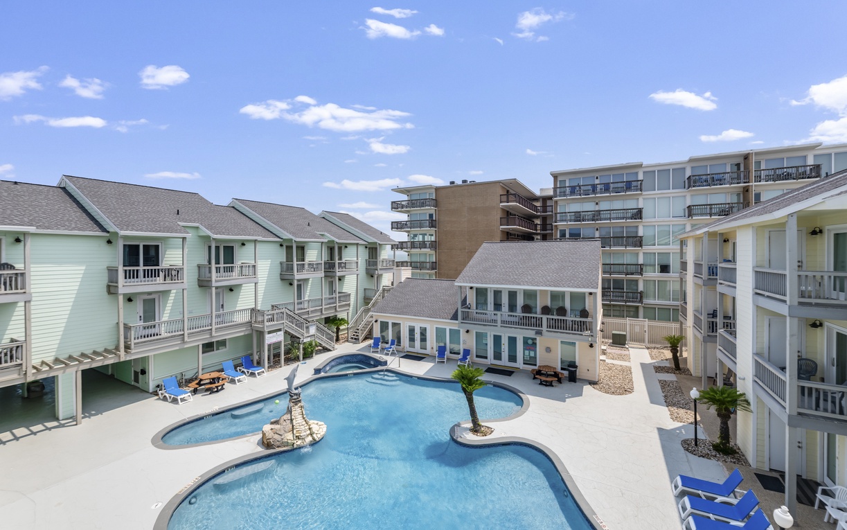 A bird's-eye view of the property reveals a beautiful resort-style pool surrounded by cheerful vacation buildings, all under a sunny coastal sky.