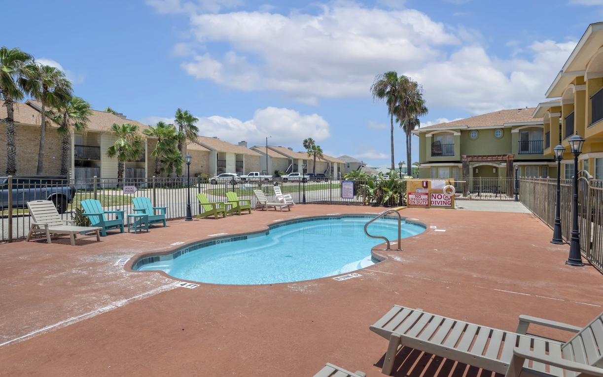 A tranquil outdoor pool area featuring palm trees, loungers in cheerful colors, and a sunny communal space perfect for relaxation and poolside leisure.