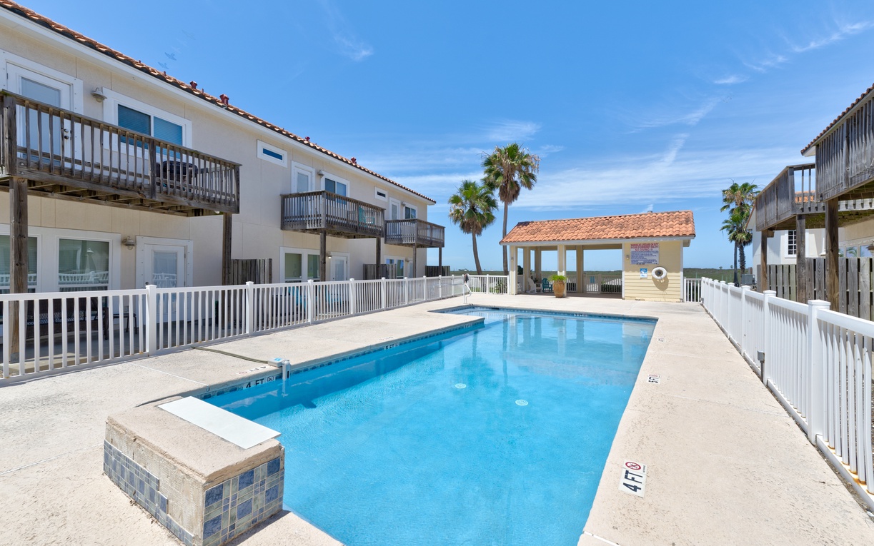 A coastal vacation rental property featuring a private pool area bordered by palm trees, with guest accommodations and a covered pavilion providing shade near the beachfront.