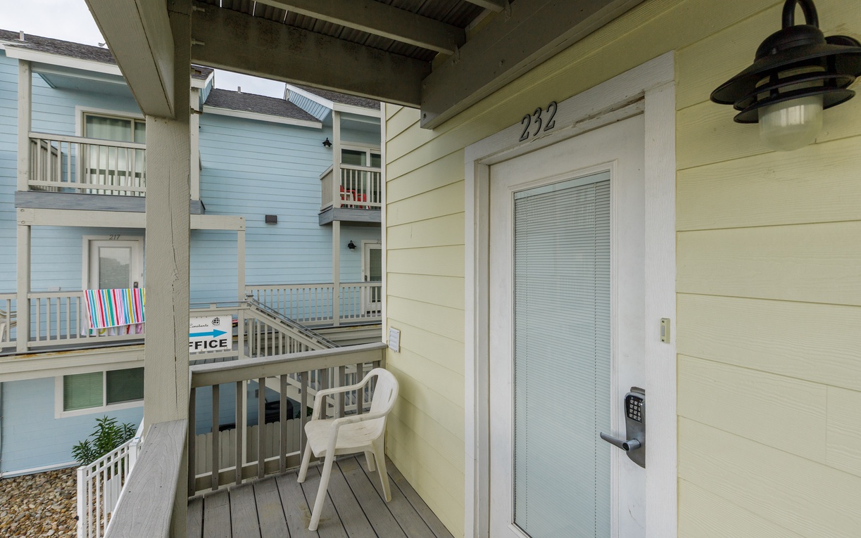 Unit 232 entrance with private balcony featuring white plastic chair and coastal neighborhood views.