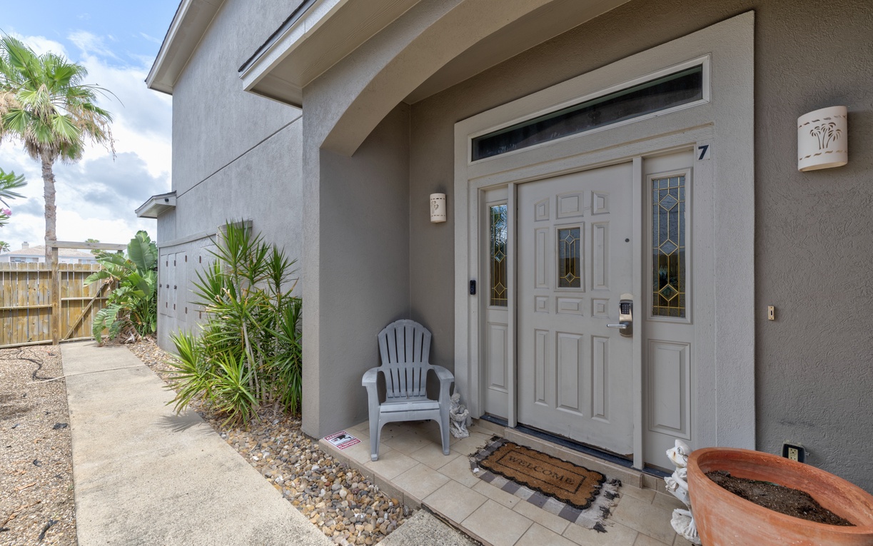 The welcoming entrance features a covered archway with decorative glass accents, tropical landscaping with palms, and a convenient smart lock for easy check-in.