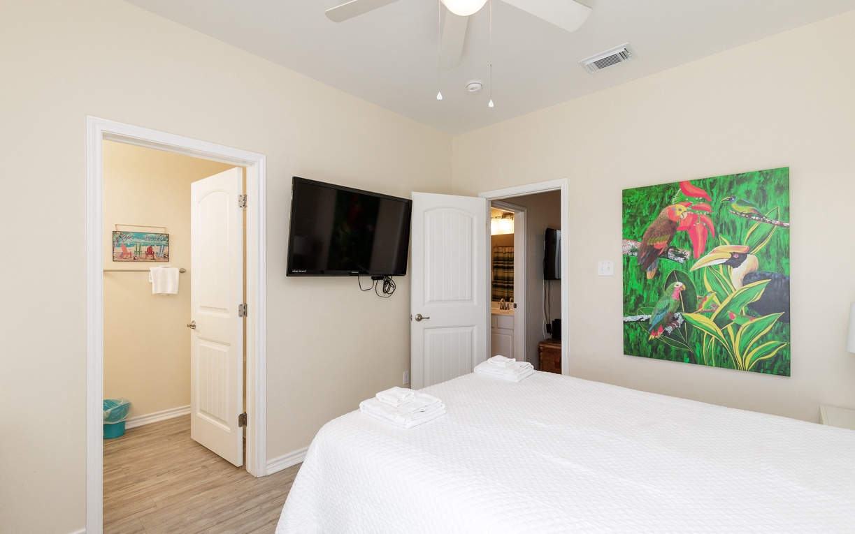 Unwind in your bright bedroom with crisp linens, ceiling fan, wall-mounted TV, and vibrant tropical artwork creating a cheerful retreat.